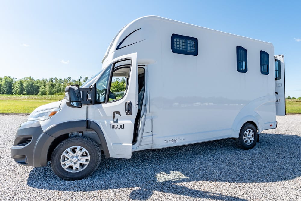 White motorhome with a raised roof and an open front door parked on gravel, Theault branding on the door side