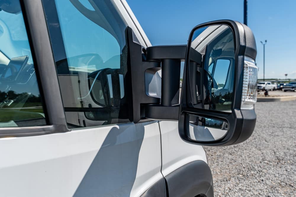 Close-up of a large side-view mirror on a white commercial truck, with a gravel lot and blue sky in the background.
