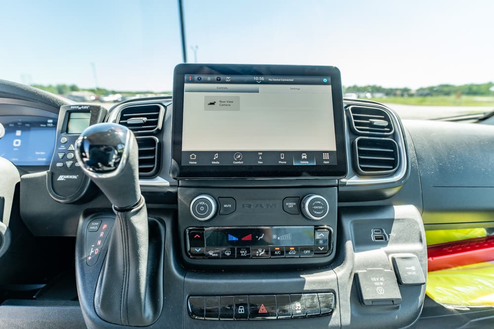 Close-up of a pickup truck cockpit showing a large center touchscreen, climate controls, and a gear shifter on the console. Sunlight streams through the windshield into the interior.