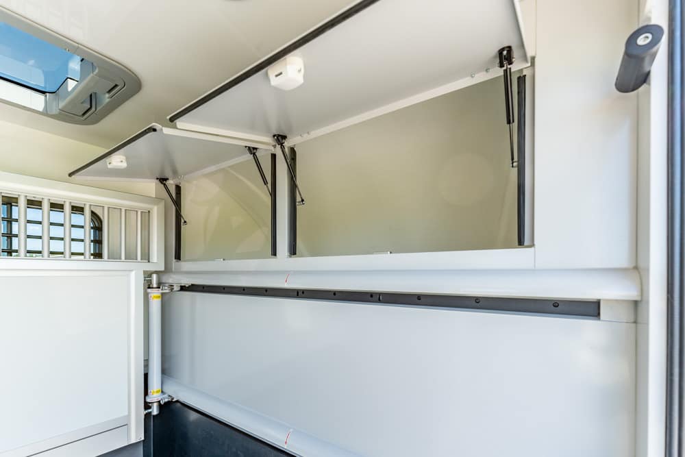 Interior of a light-gray horse trailer with upper and lower storage compartments, doors raised by gas struts, and a vented window on the left.