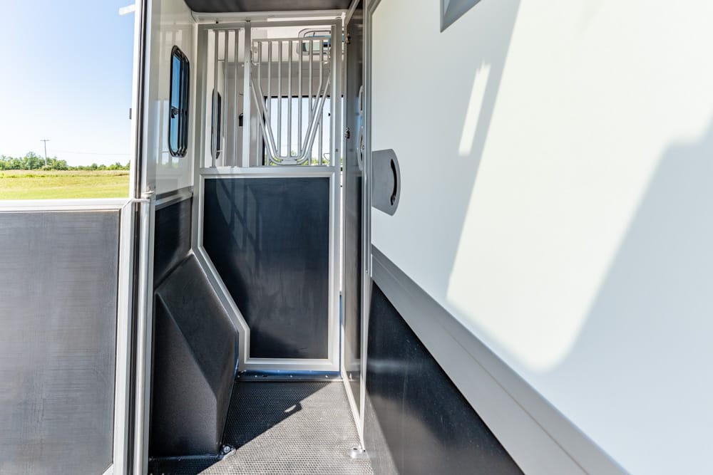 Interior of a horse trailer with metal divider bars and a partitioned stall, viewed from the open doorway toward the back on a sunny day