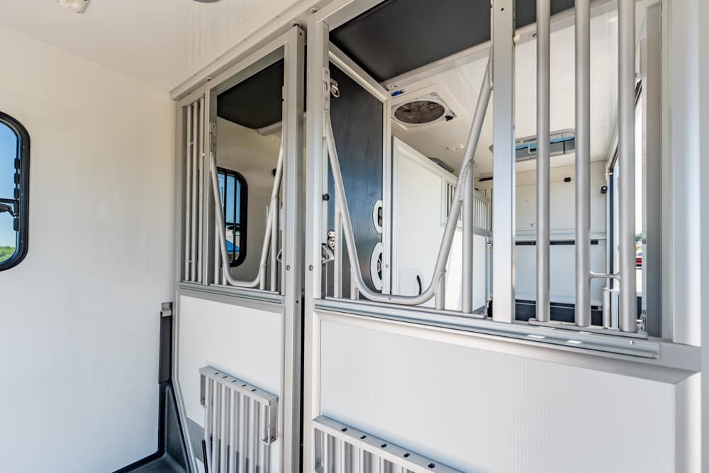 Interior of a horse trailer with metal stall dividers and closed doors, white walls visible throughout.
