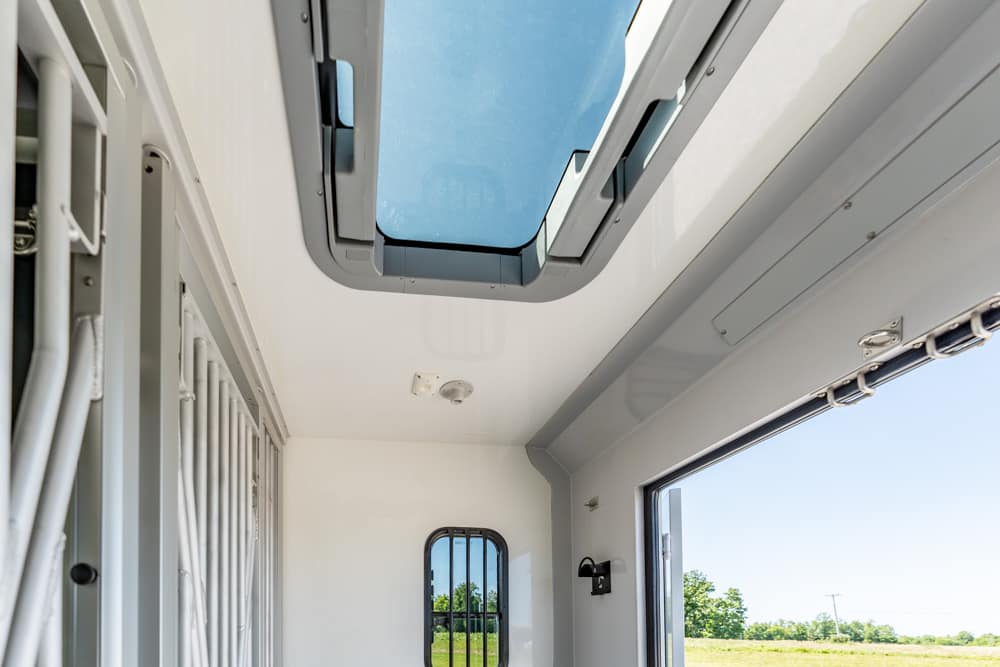 Interior of a light-gray transport vehicle with an open roof hatch and a barred window at the far end to the outside field.