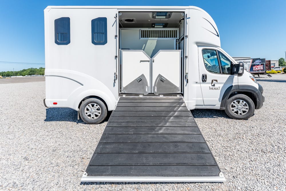 White horse trailer with the rear ramp lowered, showing two stalls inside on a gravel lot at blue-sky day