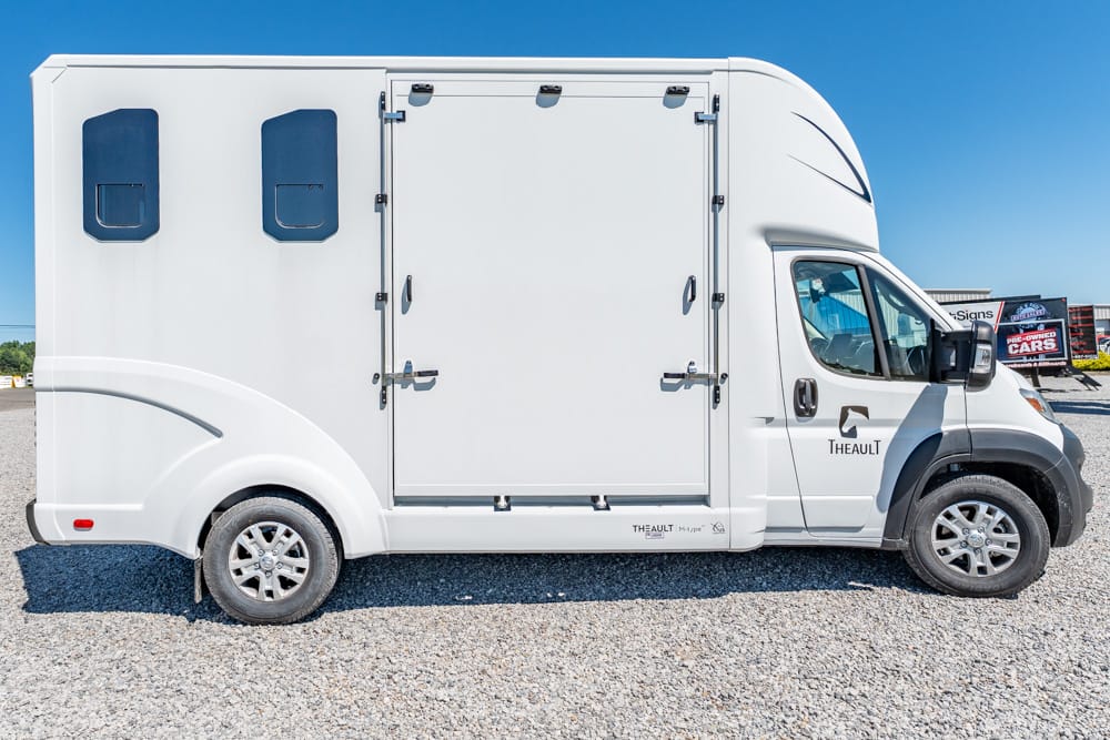 White motorhome with a large side storage compartment, two blue-tinted windows, and a cab-forward design on a gravel lot.