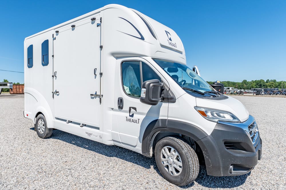 White camper van-style RV with a large box and 'THEAULT' logo, parked on a gravel lot under a blue sky (side/front view).