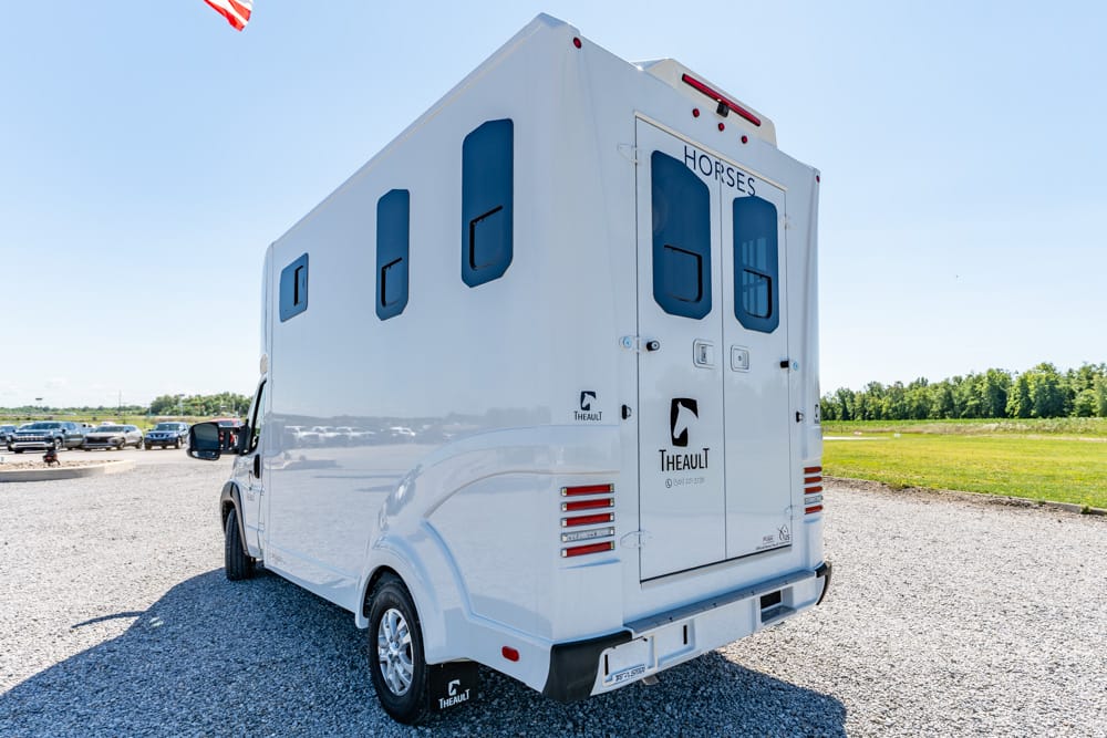 White horse-transport van viewed from the rear on a gravel lot; back doors read HORSES with Theault logo and blue-tinted windows.