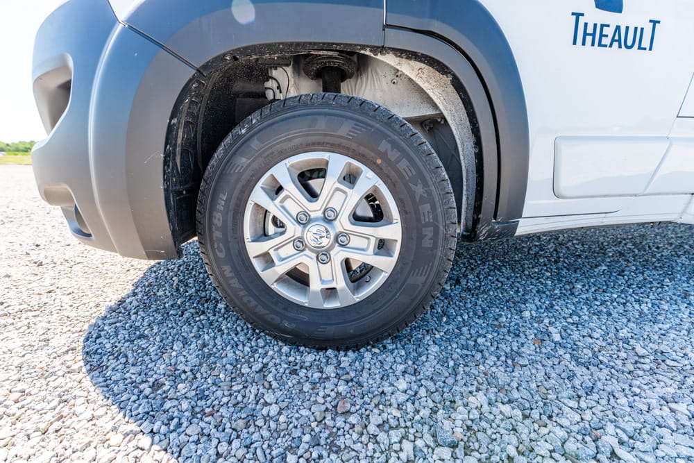 Close-up of a van's front wheel on gravel, showing the alloy rim and treaded tire with part of the wheel arch visible and Theault logo on the body.