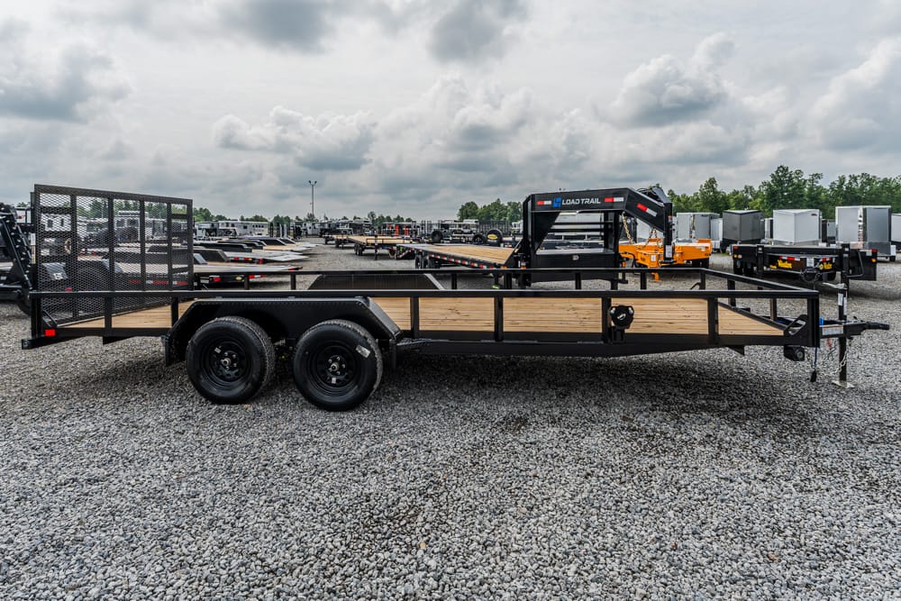 Flatbed utility trailer with wooden deck and metal railing, parked on a gravel lot among other trailers in the background.
