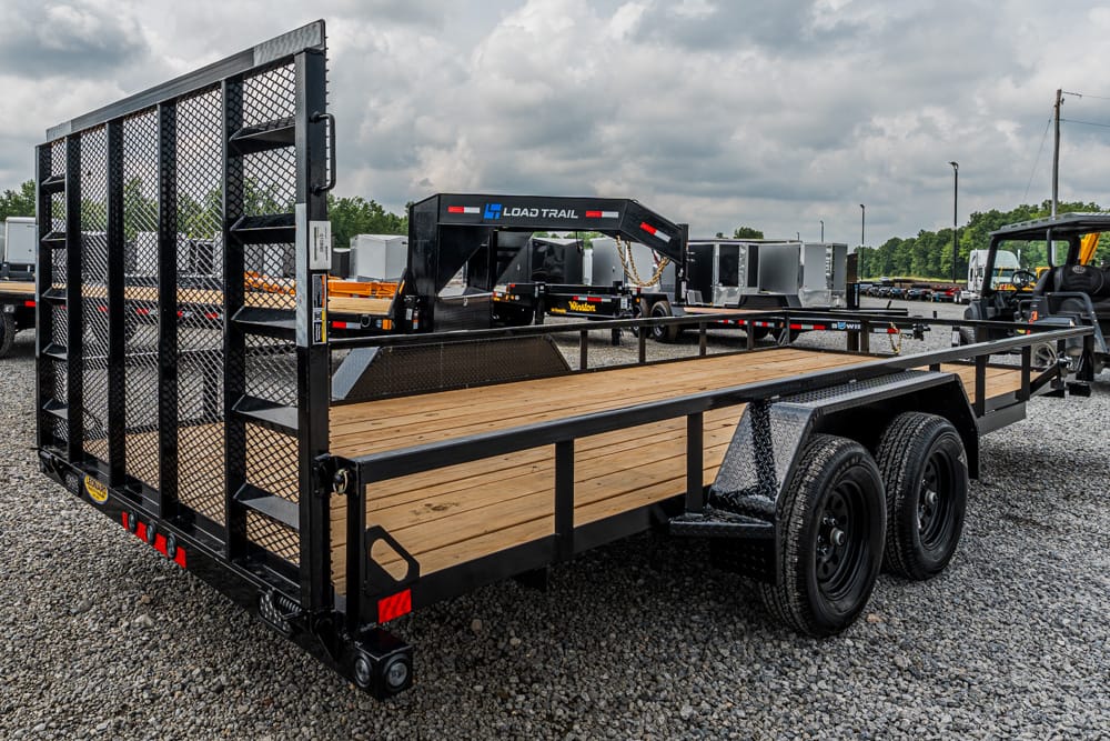 Black two-axle flatbed trailer with a wooden deck and folded mesh loading ramp at the rear, parked on a gravel lot with other trailers nearby.
