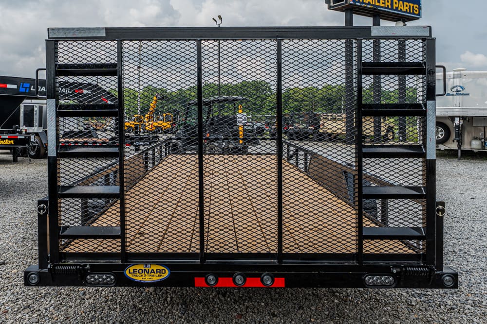 Rear view of a black utility trailer with a wooden deck and mesh sides, featuring side stairs on a gravel lot.