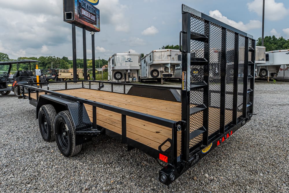 Black dual-axle utility trailer with a wood deck and a tall mesh ramp gate, parked on a gravel lot.