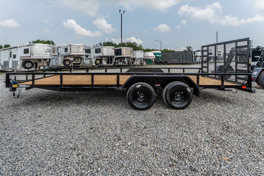 Flatbed trailer with a wooden deck and dual axles parked on a gravel lot, with horse trailers in the background.