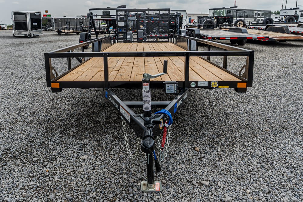 Front view of a black metal-framed utility trailer with a wooden deck, hitch and safety chains on a gravel lot.