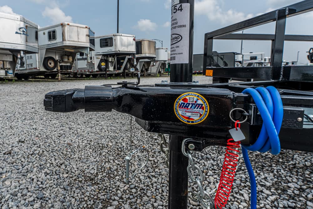 Close-up of a black trailer hitch with a blue electrical cord coiled around it on a gravel lot, with RVs in the background.