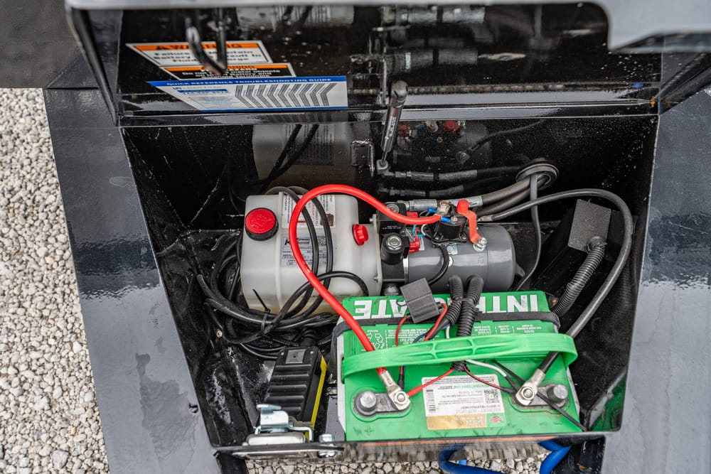 Open compartment showing a green battery with red and black cables connected to a small engine and other electrical components inside a black metal housing.