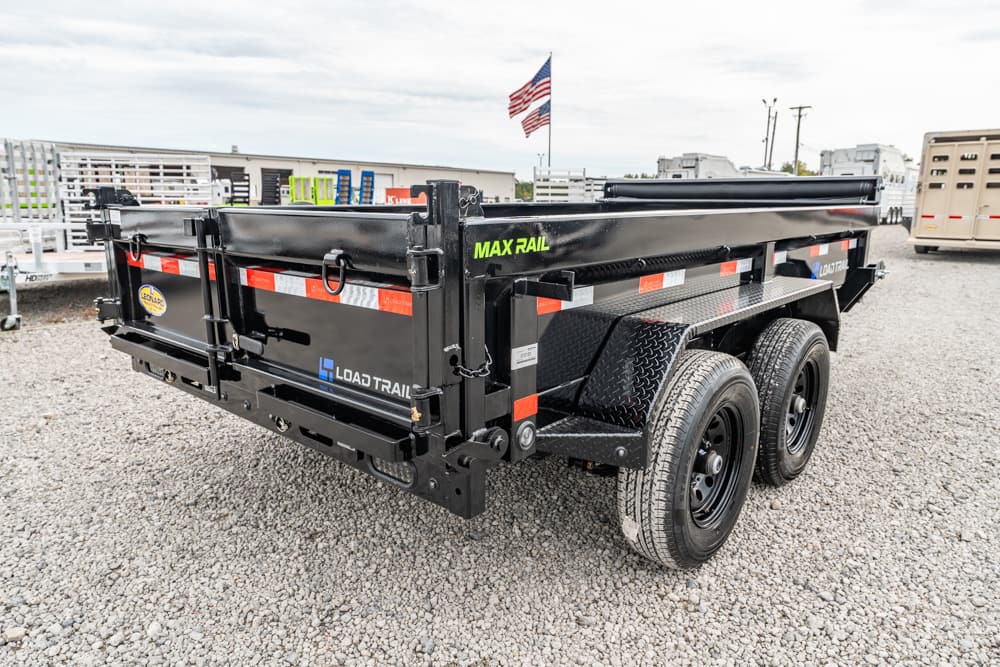 Black dual-axle Load Trail Max Rail dump trailer on a gravel lot with an American flag in the background.