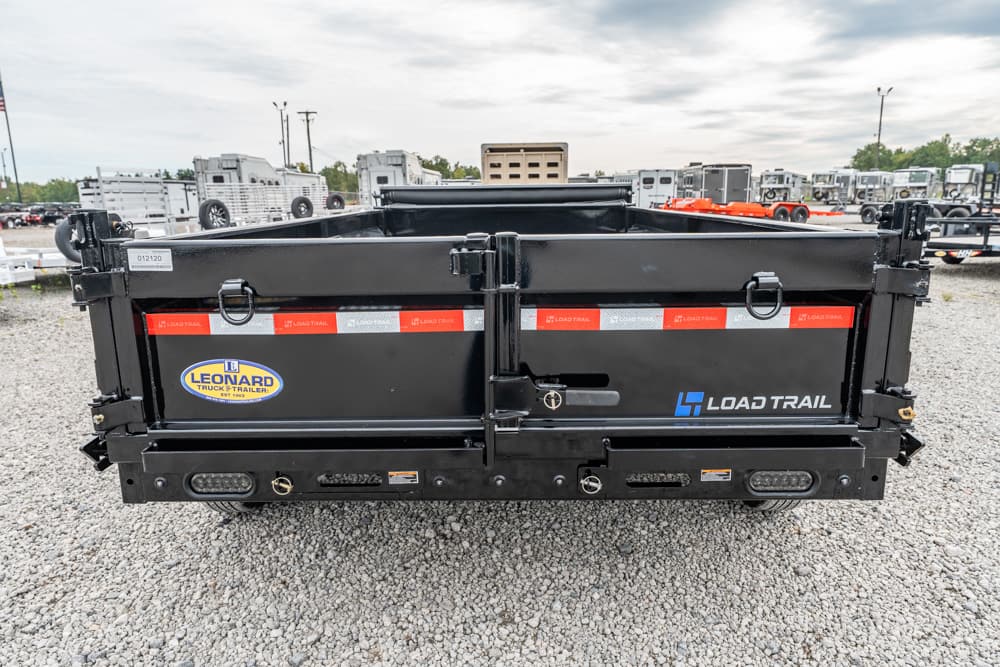 Rear view of a black Load Trail dump trailer with reflective red-white stripes and Leonard sticker in a gravel lot.