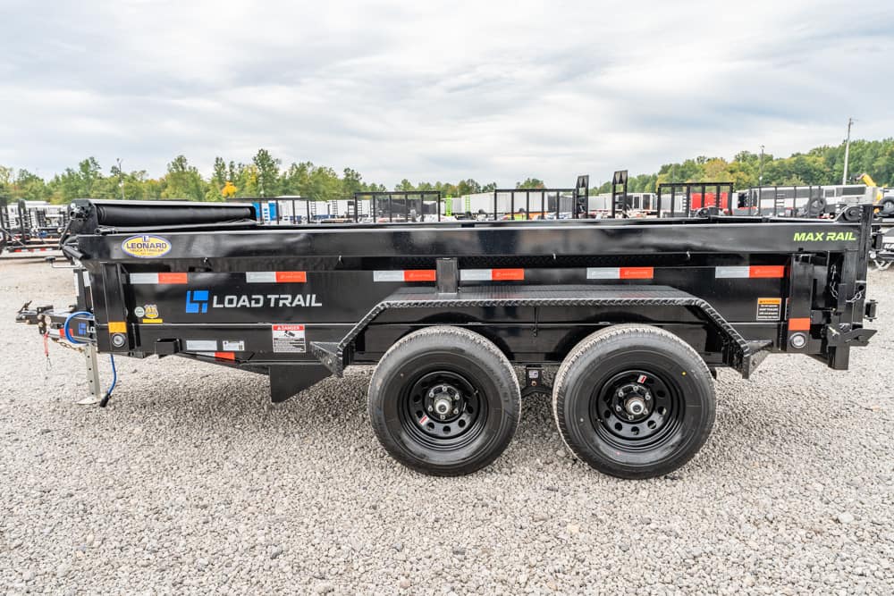 Side view of a black Load Trail Max Rail dump trailer with dual axles parked on gravel, showing red reflectors and brand logos on the side