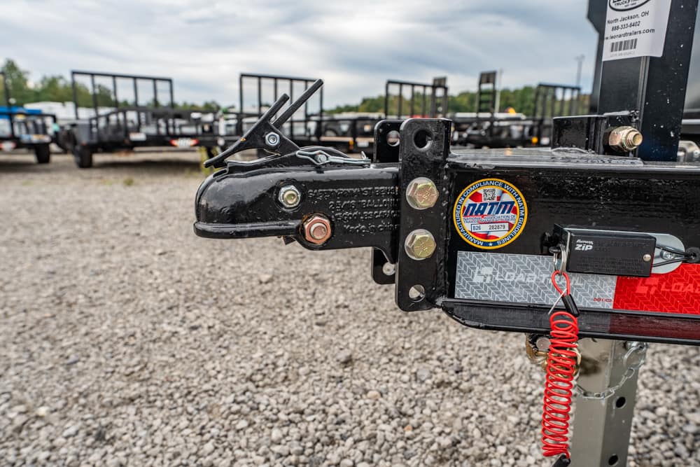 Close-up of a trailer hitch on a gravel lot, showing a safety latch, bolts, and a red coiled safety cord with labels nearby
