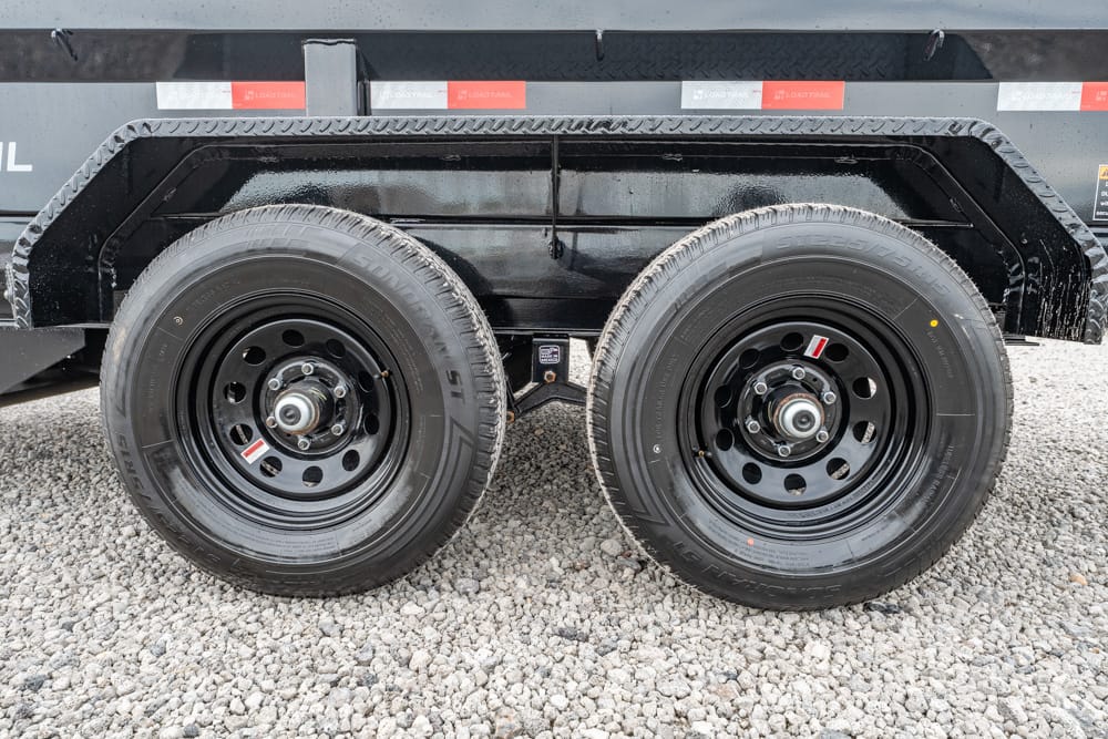 Close-up of a tandem trailer wheel assembly with black rims and tires on a gravel surface under a trailer bed.