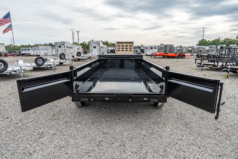 Black flatbed trailer with side gates opened wide, parked on a gravel lot with other trailers in the background.