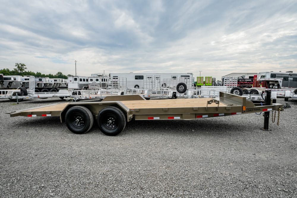 Flatbed trailer with dual axles and a wooden deck parked on a gravel lot with other trailers in the background, showing utility equipment ready for sale or rent.