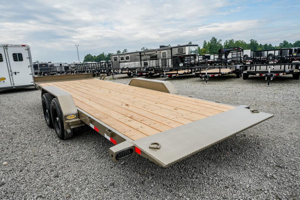 Flatbed trailer with a wooden deck and lowered rear ramp on a gravel lot, surrounded by other trailers and equipment.