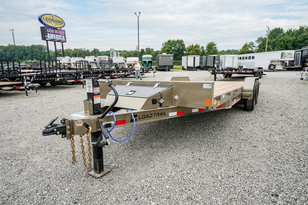 A silver Load Trail flatbed trailer on a gravel lot, with blue tow hitch and safety chains in the foreground, and other trailers in the background.