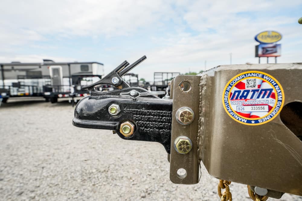 Close-up of a trailer hitch coupler with bolts and a safety latch, attached to a brown frame with a DOT compliance sticker in view behind it.