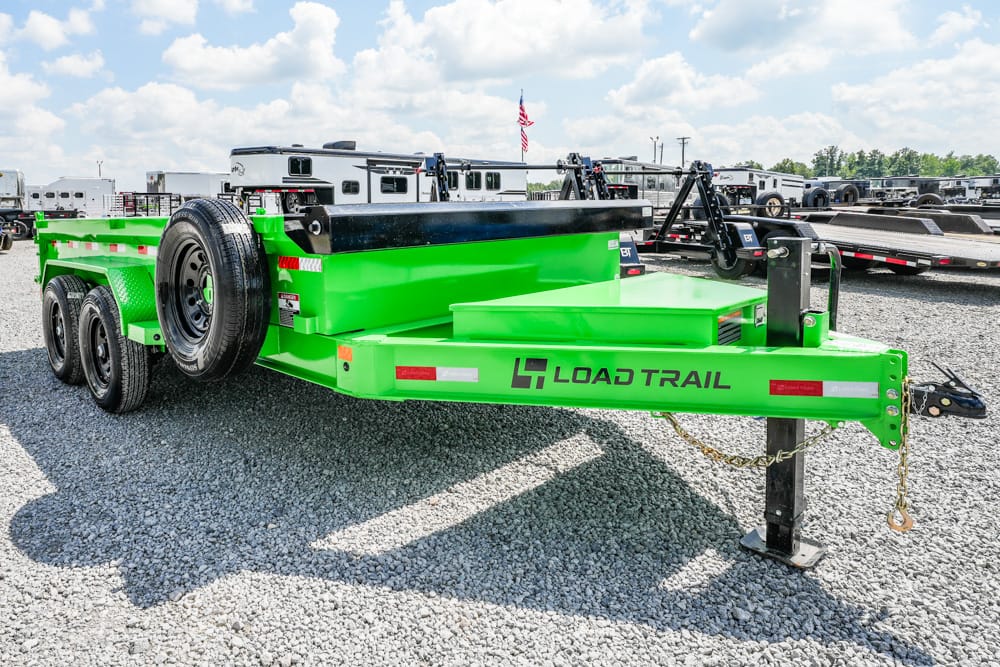 Bright green Load Trail equipment trailer parked on gravel, with spare tire mounted on the side and other trailers in the background.