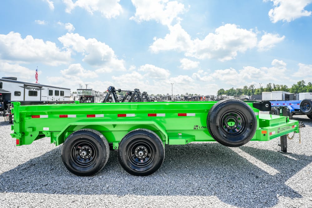 Bright green multi-axle trailer with two rear axles and a spare tire mounted on the side, parked on a gravel lot with other trailers in the background