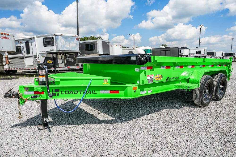 Bright green Load Trail tandem-axle dump trailer on a gravel lot, with a hitch jack and blue interaction hoses, and several trailers in the background.