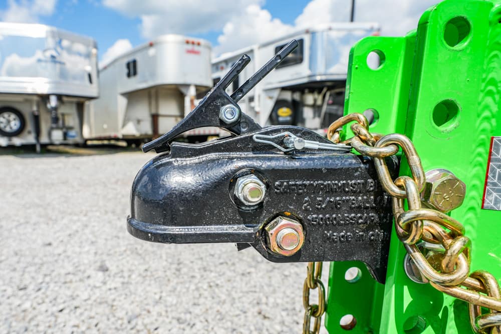 Close-up of a black trailer hitch with a safety chain attached to a green trailer frame, outdoor lot in background.