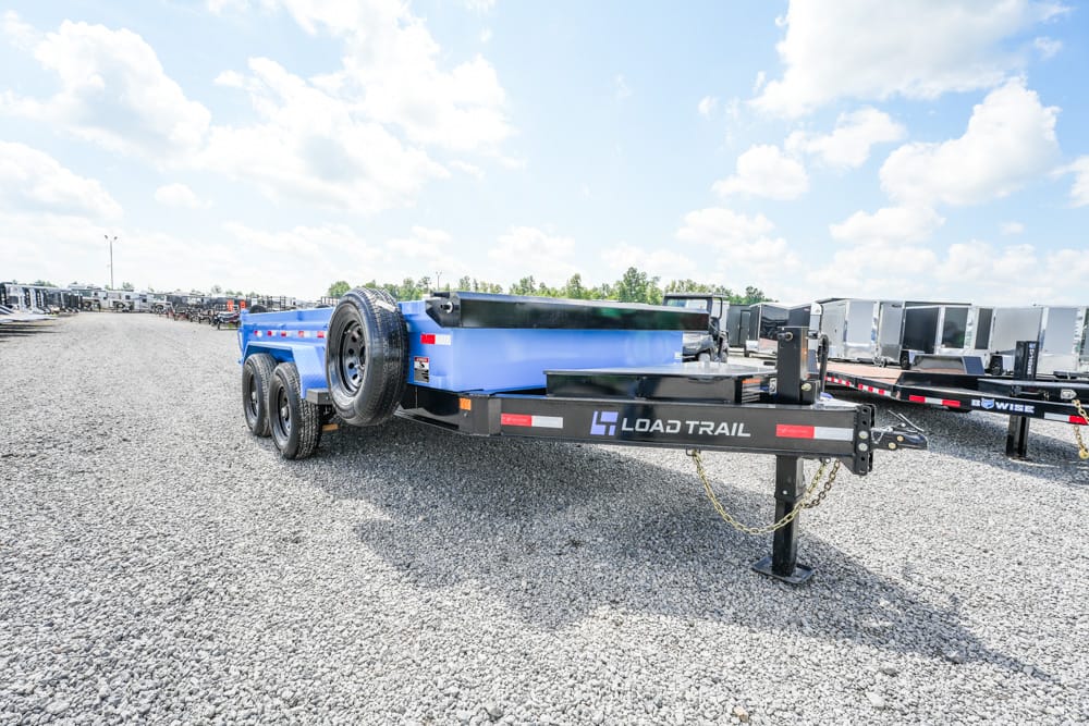 Blue Load Trail utility trailer on a gravel lot, dual-axle with a spare tire mounted on the front, surrounded by other trailers under a bright, partly cloudy sky.
