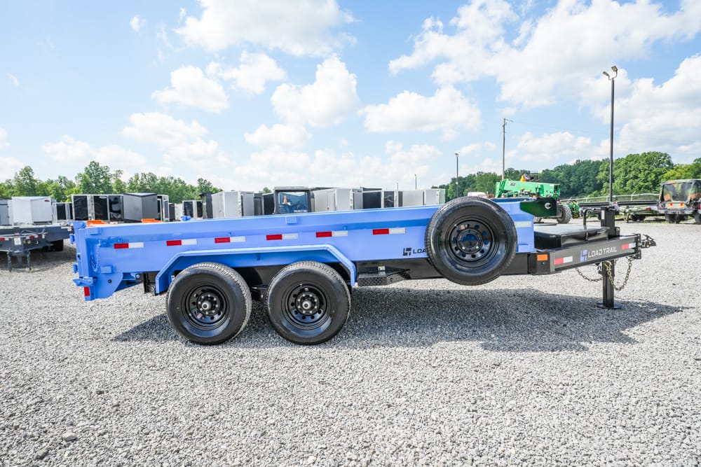 Blue flatbed trailer with three axles and a side-mounted spare tire, parked on a gravel lot with other trailers in the background.