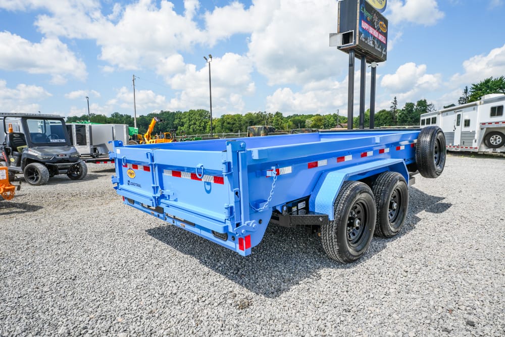 Blue tandem-axle dump trailer on a gravel lot, with a spare tire mounted on the side and other equipment in the background.