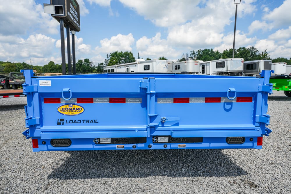 Rear view of a bright blue Load Trail dump trailer with safety latches and red reflectors in a gravel lot.