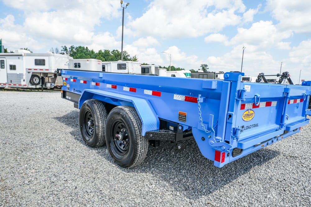 Blue heavy-duty dump trailer with dual rear axles parked on gravel lot, surrounded by RVs and trailers under a partly cloudy sky. Intended for hauling materials; shows safety chains and reflective strips.