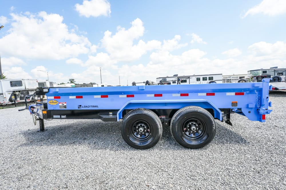 Blue Load Trail dump trailer with dual axles parked on gravel lot; several trailers/RVs visible in background.