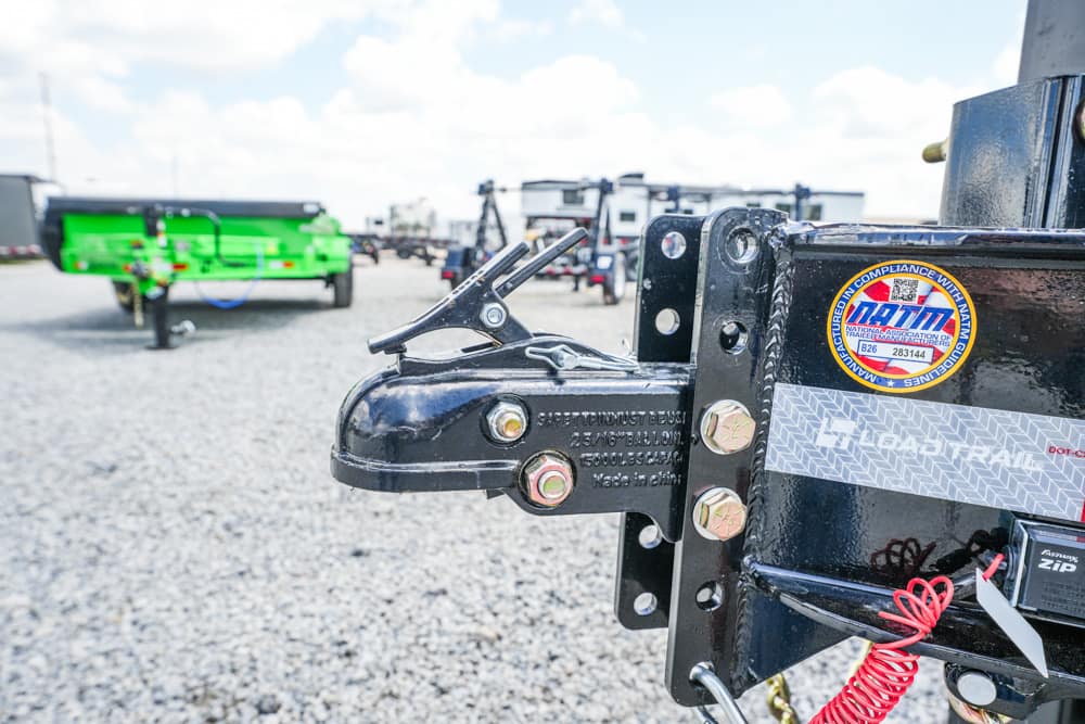 Close-up of a black trailer hitch latch and coupler with bolts, a gravel lot and a bright green trailer in the background.