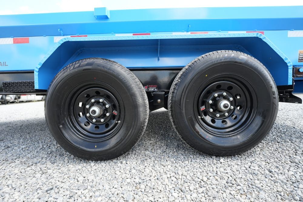 Blue cargo trailer side with dual rear wheels on a gravel surface, black rims and tire treads visible in daylight