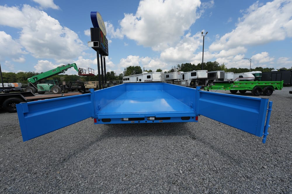 Blue dump trailer with lowered sidewalls viewed from the rear in a gravel lot, ready to haul material; other equipment and trailers in the background.