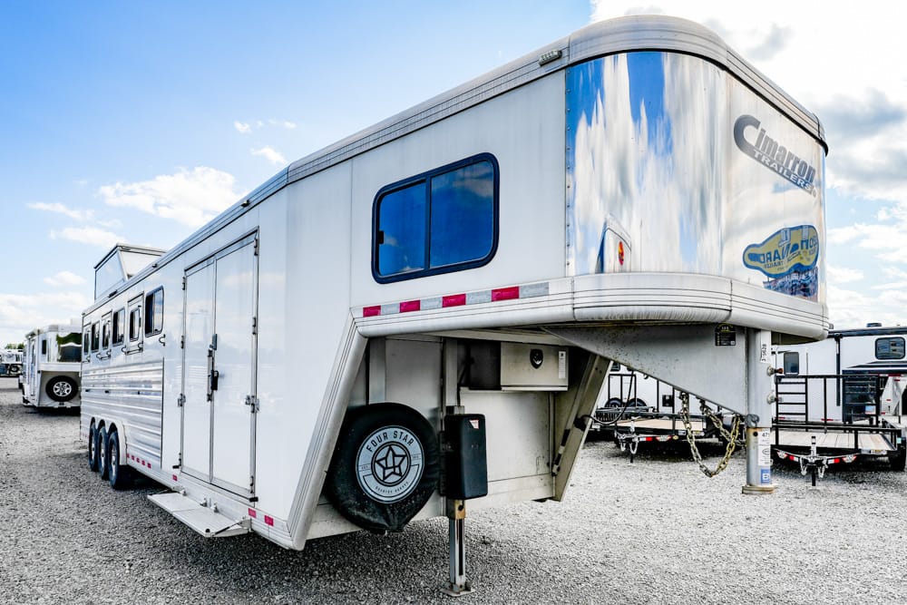 White gooseneck horse trailer parked on a gravel lot, spare tire mounted on the side and windows along the length.