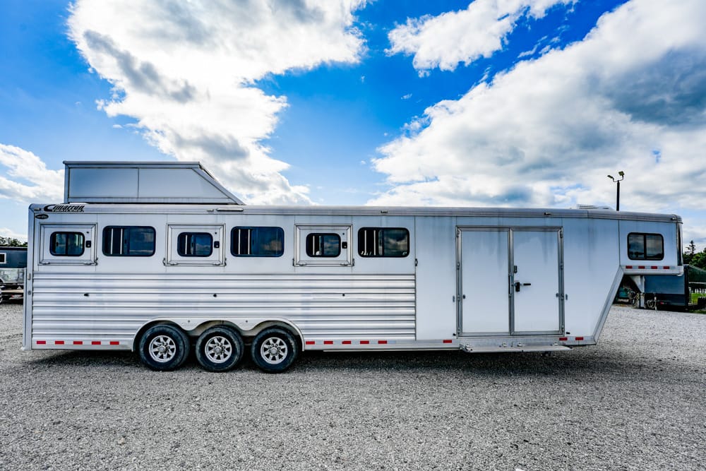 Large white horse trailer parked on a gravel lot under a blue sky with fluffy clouds.