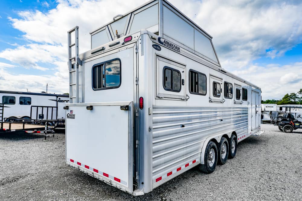 White horse trailer with multiple side windows and a rear ramp, parked on a gravel lot under a partly cloudy sky.