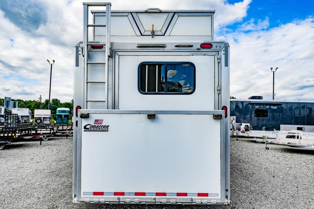 White horse trailer seen from the rear with a small window, ladder on the left, and other trailers on a gravel lot under a partly cloudy sky.
