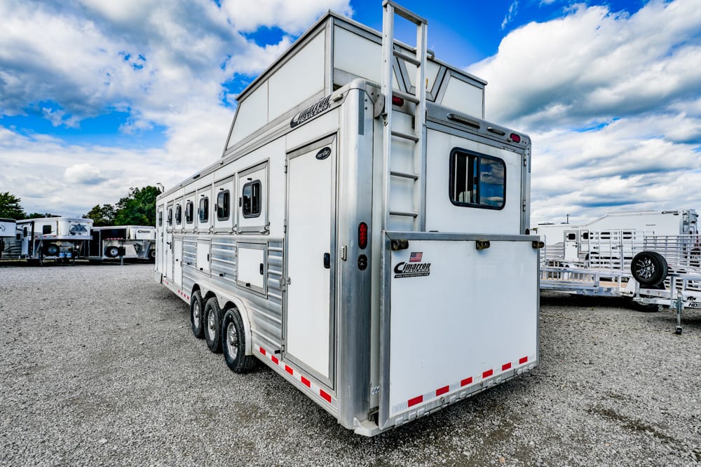Rear and side view of a triple-axle white aluminum horse trailer with a back ladder and Cimarron branding, parked on a gravel lot with other trailers nearby.