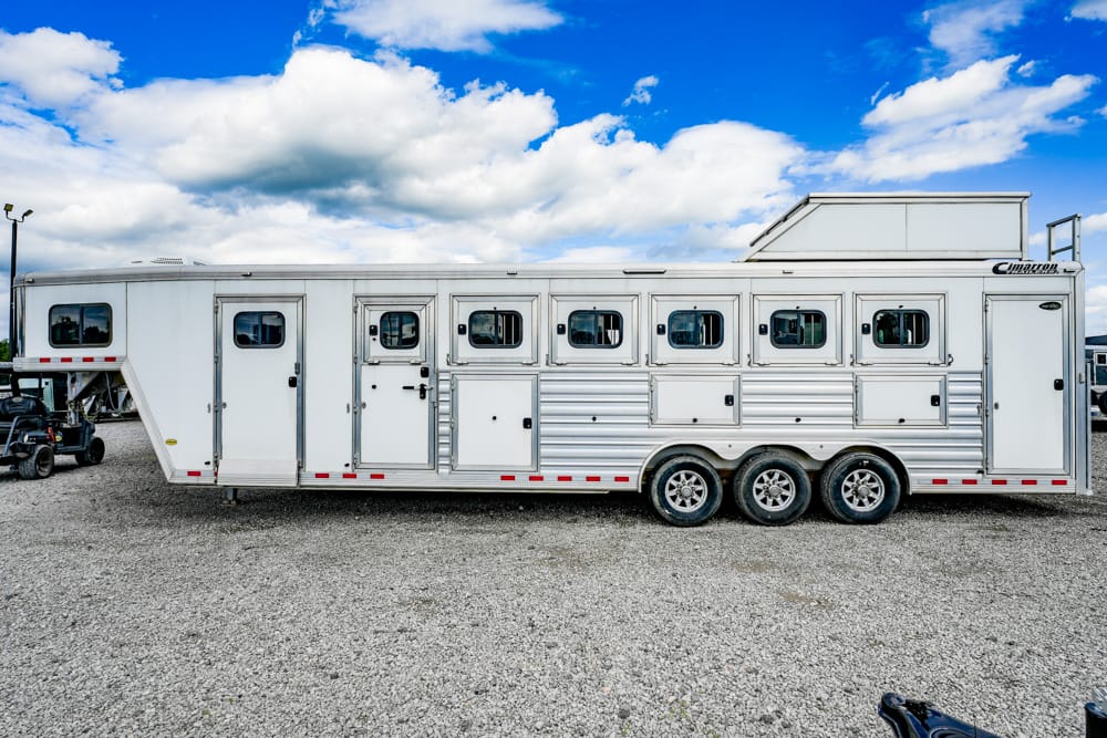 White long horse trailer with multiple windows and three axles parked on a gravel lot under a blue sky with clouds, side view