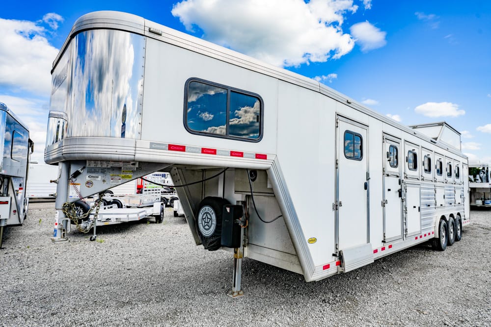 Large white gooseneck horse trailer on a gravel lot, featuring multiple windows, storage compartments, and a spare tire underneath, under a blue sky.